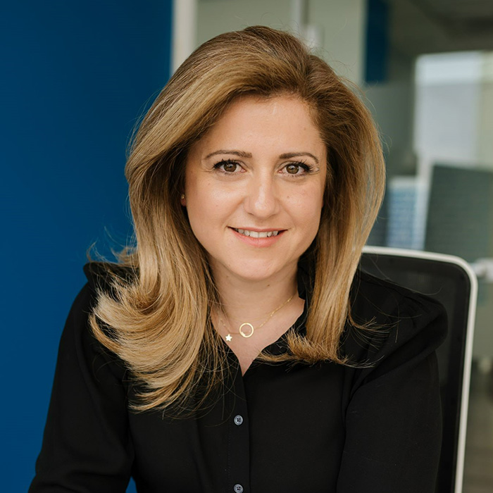 A woman smiles slightly towards the camera as she sits on a chair in an office space. She has a loose hairstyle and is wearing a black shirt.