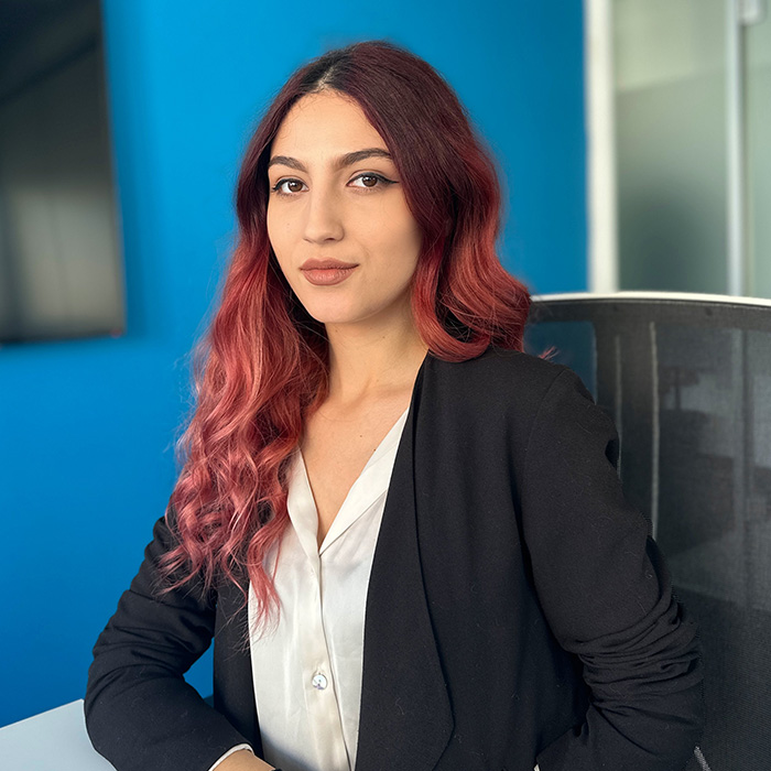 A woman smiles slightly towards the camera as she sits on a chair in an office space. She has a loose hairstyle and is wearing a black shirt.