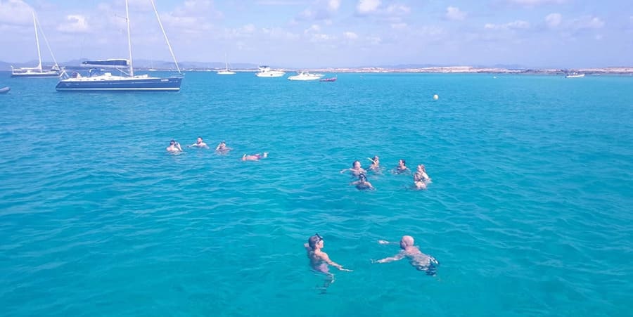 Un grup de oameni se bucură de o baie în ocean. În depărtare se observă ambarcațiuni.
